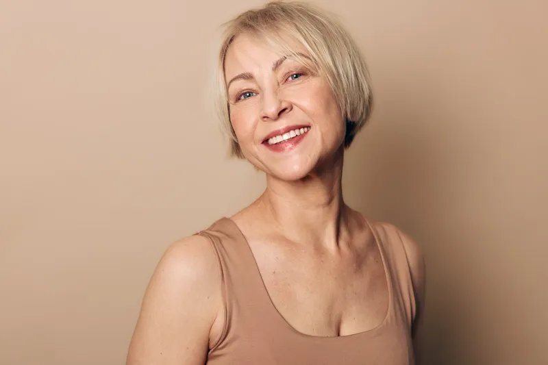 Smiling middle aged woman with short blonde hair in beige tank top looks happy confident on plain background. Concept of natural beauty, aging gracefully, wellness and positive mood.