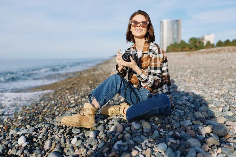 Photographer woman sits on a pebble beach with a camera, wearing a plaid shirt and boots, capturing a coastal scene under bright sun, conveying relaxed outdoor lifestyle and creative travel moments