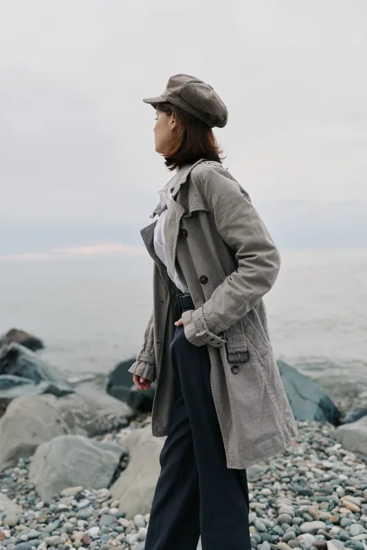 Young woman in stylish coat and cap stands on rocky shore looking at calm sea under cloudy sky. Concept of autumn fashion, outdoor lifestyle, and contemplative mood.