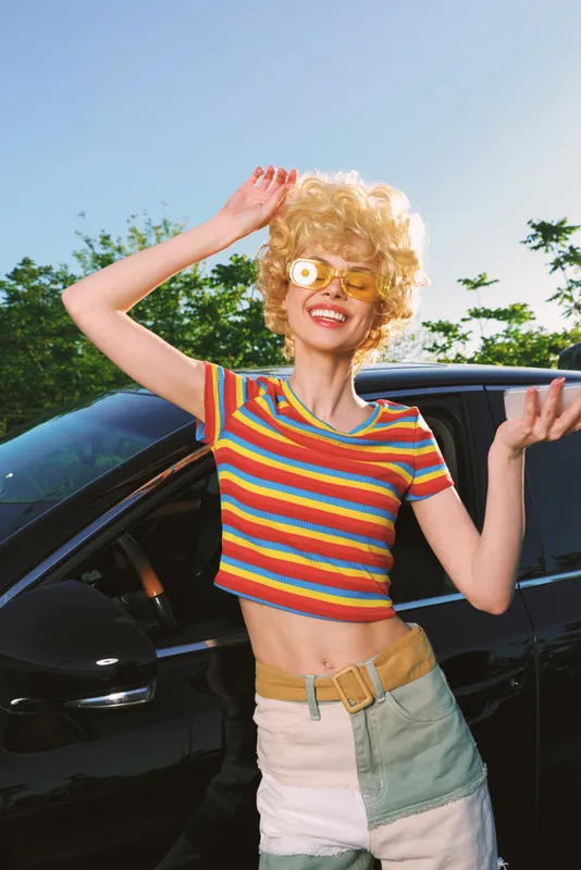 Colorful retro woman poses beside a sleek car, smiling behind flower sunglasses, wearing a striped crop top, sunny day, carefree lifestyle shot with vibrant vintage film color and glow.