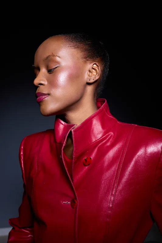 Elegant black woman with dark skin wears a shiny red leather jacket, posing with eyes closed against a dark background. Concept of fashion, confidence, and beauty portrait.