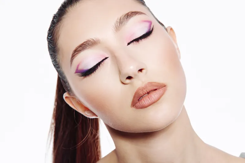 Young woman with unusual creative eye makeup and nude lipstick poses with closed eyes. The image focuses on artistic beauty and modern fashion. Clean white background.