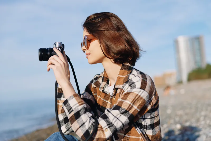 Photographer, camera in hand, woman studies the shoreline in a plaid jacket, sunglasses, and calm focus as waves roll in and distant buildings frame a relaxed outdoor scene.
