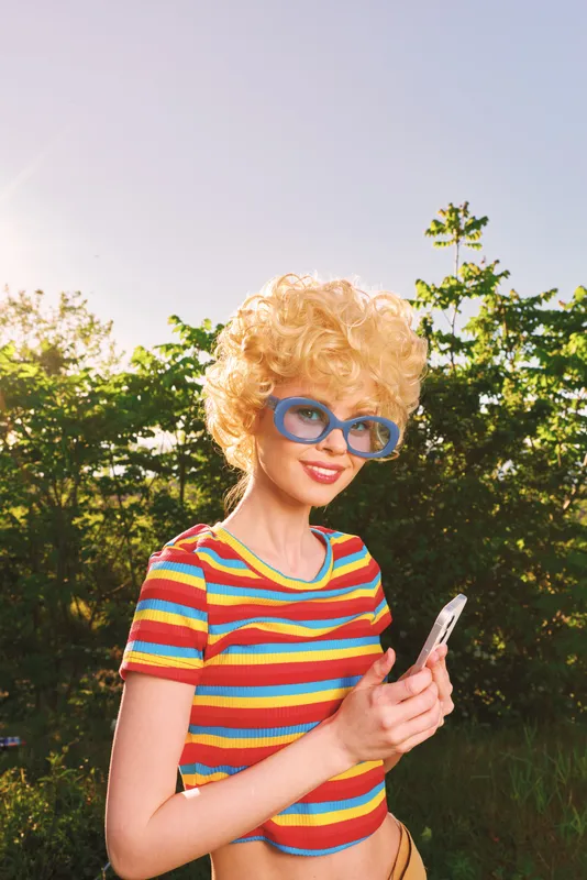 A playful retro woman outdoors wears a colorful striped shirt and oversized blue sunglasses, smiling while checking her phone. A lighthearted lifestyle moment with vintage flair and sunny mood.