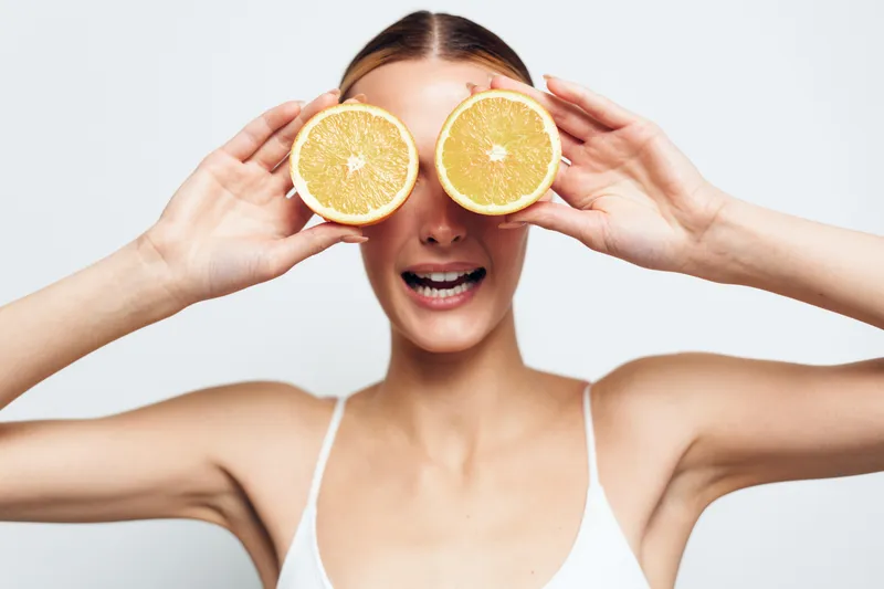 Young woman in white tank top holds two orange halves over her eyes while smiling playfully against a plain light background.