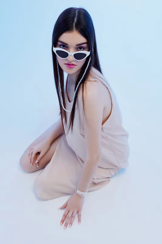 Young woman in beige outfit and white sunglasses poses sitting on white floor. Her long dark hair and stylish look highlight fashion and beauty concepts in studio lighting.