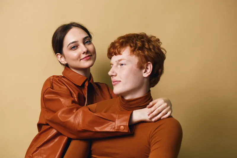Couple embracing and smiling, young woman and man in warm clothing looking happy together, posing in studio with beige background and casual stylish outfits.