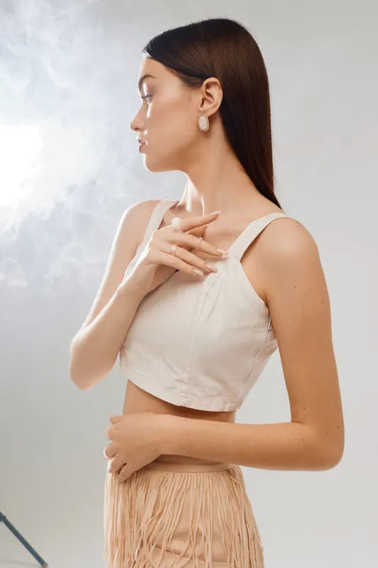 Young woman in white top and beige skirt poses sideways with hand near chest. Soft lighting highlights her smooth skin and elegant earrings. Concept of beauty fashion and style.