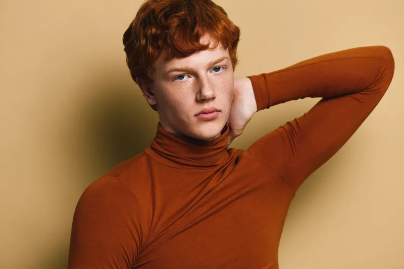Young man with red hair wearing brown turtleneck posing with hand behind head against beige background looking confident and thoughtful in studio portrait with soft lighting.