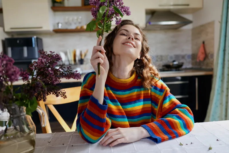 Cheerful woman indoors, wearing a colorful striped sweater, sits at a table with flowers, bright kitchen in background, relaxed mood, soft light, candid lifestyle moment of everyday joy