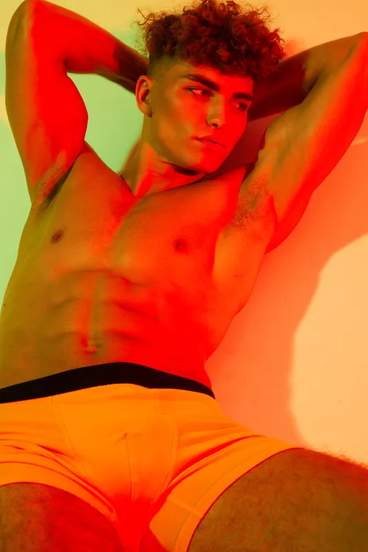 Young man with muscular body posing shirtless in bright orange shorts against colorful background. Fitness, model, toned physique and confident expression in studio lighting setup.