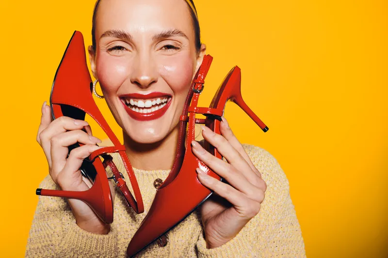 Happy woman with bright red lipstick holds three red high heel shoes close to her face against vibrant yellow background. Concept for fashion, style, and joyful expression.