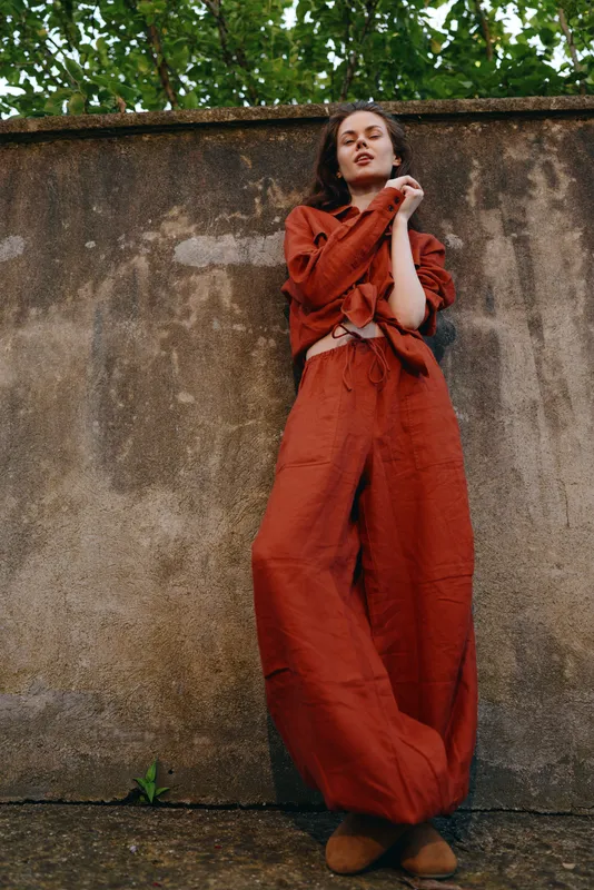 A woman in a rust red jumpsuit leans against a weathered wall, arms crossed, eyes closed, conveying calm confidence. A lifestyle moment blending fashion, comfort, and urban outdoors, captured in