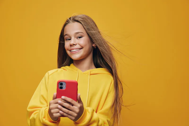 Teenage girl smiling and holding smartphone for social media and communication. Casual young female with long hair wearing yellow hoodie on yellow background with happy expression.