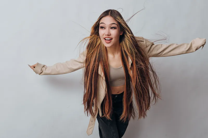 Young woman with long straight hair and casual outfit posing with arms outstretched against plain light background for fashion and lifestyle portrait.