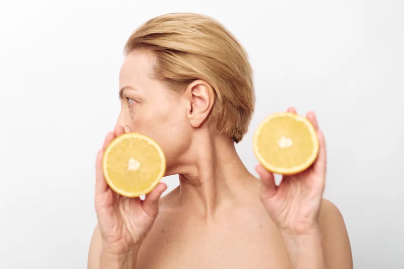 Blonde woman with short hair holds two halves of a citrus fruit near her face, looking sideways against a plain white background. Concept of natural skincare, freshness, and healthy lifestyle.