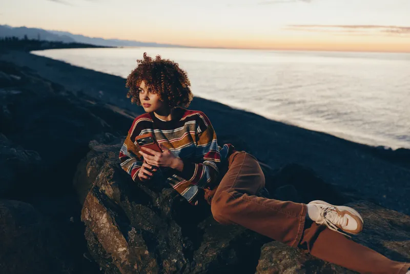 Woman lifestyle, sunset over sea and ocean, outdoors on a rock by the shore, curly hair framing her face, patterned sweater and jeans, seated and looking toward the horizon, calm