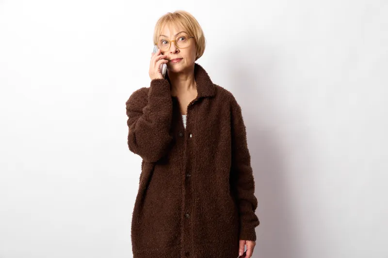 Woman senior phone call communication glasses coat white background indoors holding smartphone to ear focused expression copy space during daytime photo