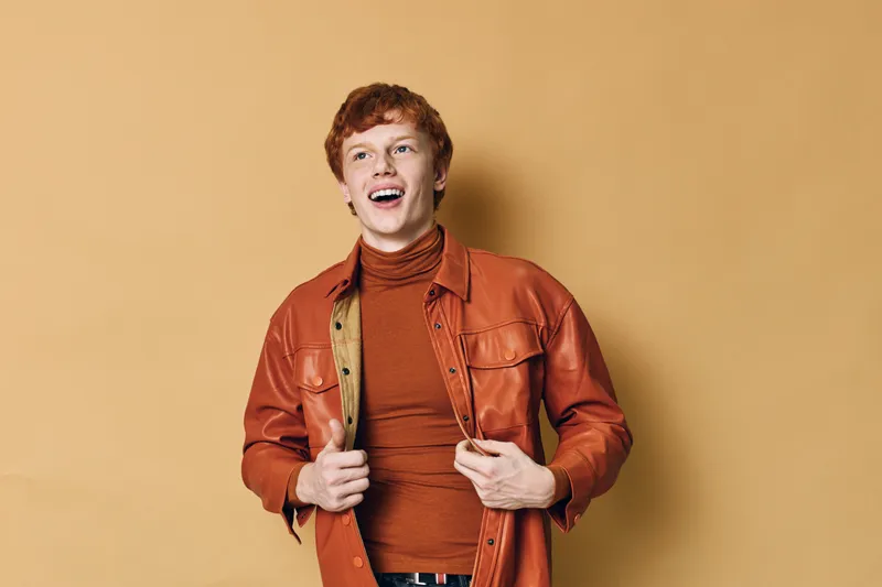 Young man with red hair wearing orange jacket and turtleneck smiling and looking away standing against solid beige background with casual style and confident expression.