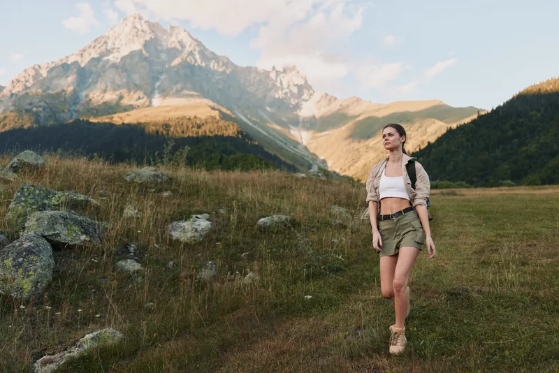 A woman hiking in a sunlit field with towering mountains in the distance, capturing outdoors lifestyle, adventure, and nature within a vivid landscape for stock use.