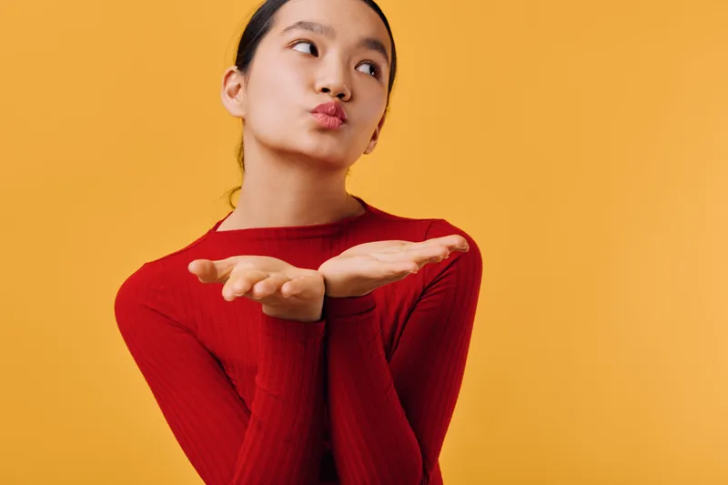 Woman with long dark hair wearing red sweater poses with hands near face blowing a kiss on yellow background. Portrait of young female sending air kiss for love or greeting.