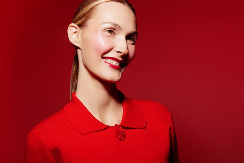Smiling woman with red lipstick and red sweater poses on red background. Bright studio lighting highlights her clear skin and joyful expression. Concept of beauty and fashion.