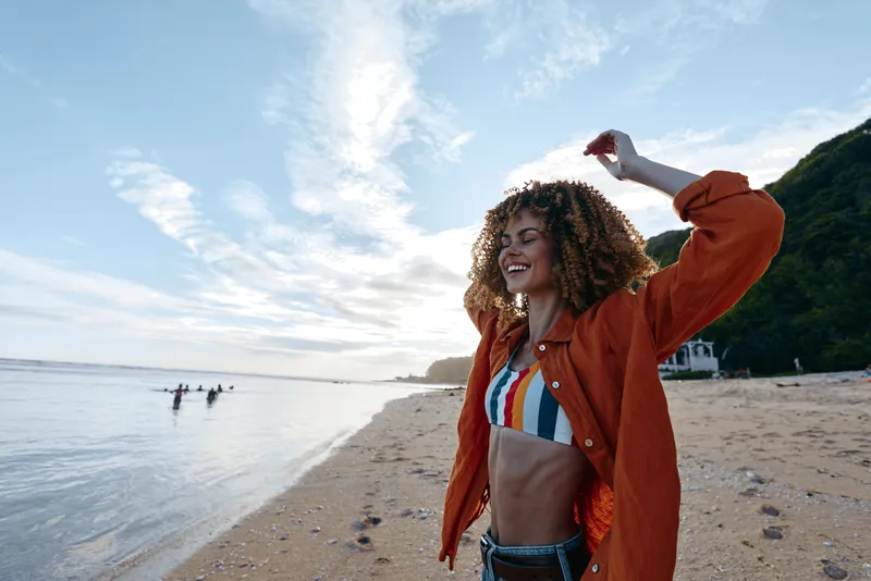 Young woman with curly hair wears a striped bikini under an orange jacket, smiling as she dances on a sunlit beach, and the scene captures carefree energy, warm light, and a sense of summer freedom.
