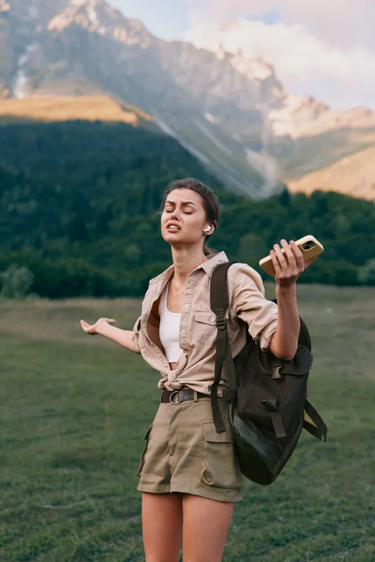 Confident woman outdoors with backpack standing in a grassy meadow mountains in the background arms open smiling conveying travel nature and self discovery