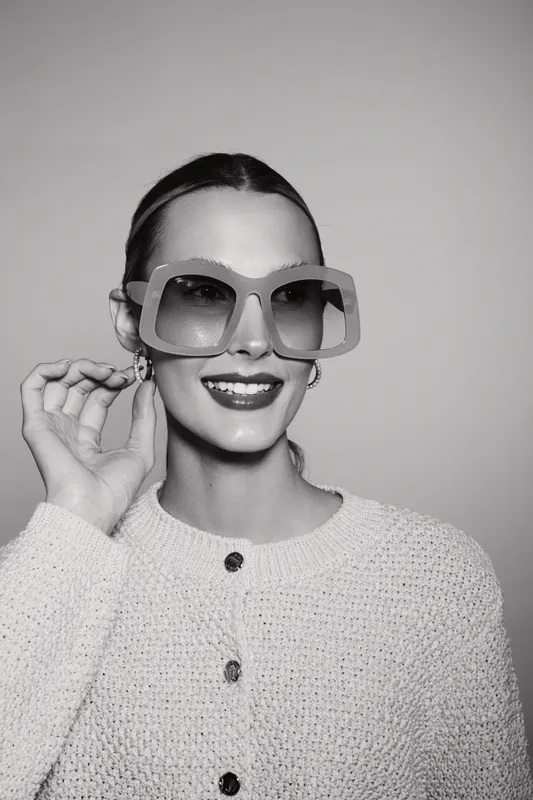 Smiling Caucasian woman with fair skin wearing oversized sunglasses and a knitted sweater poses against plain background. Concept of fashion, style, and eyewear accessories.