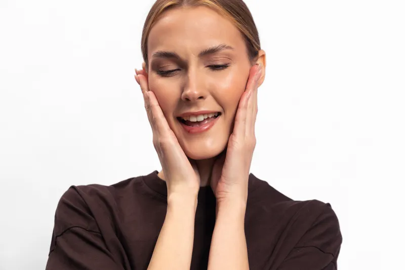 Young woman with clear skin smiles gently with eyes closed, hands touching face softly. Concept of skincare, beauty, relaxation, and natural wellness in studio setting.