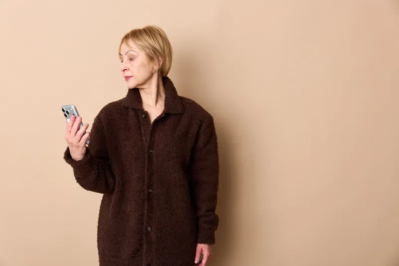 Woman holding smartphone indoors in studio, casual dark coat, communication through texting with focused expression, neutral beige background, soft light, calm modern mood