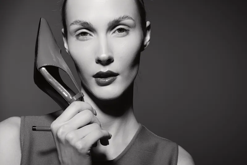 Closeup of pale white woman holding high heel shoe near face with serious expression on dark background.