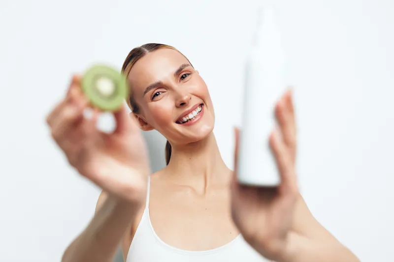Smiling woman holds kiwi fruit and white cosmetic bottle showing natural skincare concept with fresh fruit, healthy lifestyle, and beauty care on white background in studio.