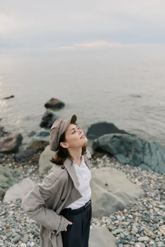 Young woman in casual outfit and hat stands on rocky beach with closed eyes, enjoying fresh air and peaceful sea view. Concept of relaxation, nature, and mindfulness outdoors.