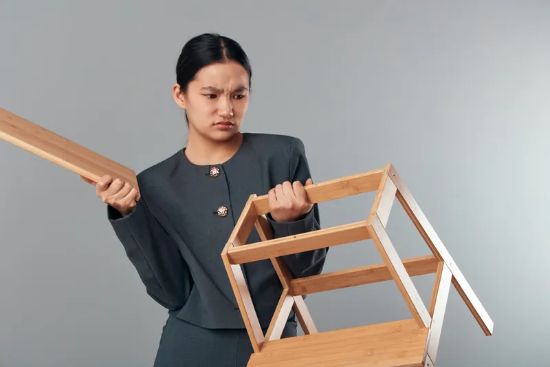 Angry woman holding wooden chair and looking frustrated indoors. Female in formal outfit with furrowed brow gripping furniture against gray background in studio. Emotion and tension.