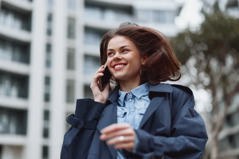 Smiling woman in a denim jacket talks on a mobile phone in an urban outdoor setting, with modern buildings in the background, bright natural light, and a confident mood.