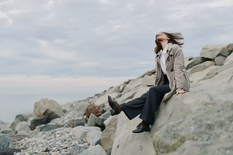 Young woman in casual outfit sits on large rocks by the sea, enjoying fresh breeze and cloudy sky. Concept of freedom, nature, relaxation, and outdoor lifestyle near water.