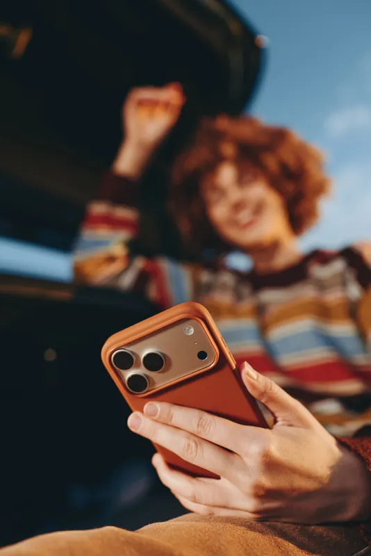 Woman lifestyle, closeup hand holds orange smartphone and phone, taking photo, blurred young woman in striped sweater, casual indoor home scene, warm playful mood, shallow depth of field.