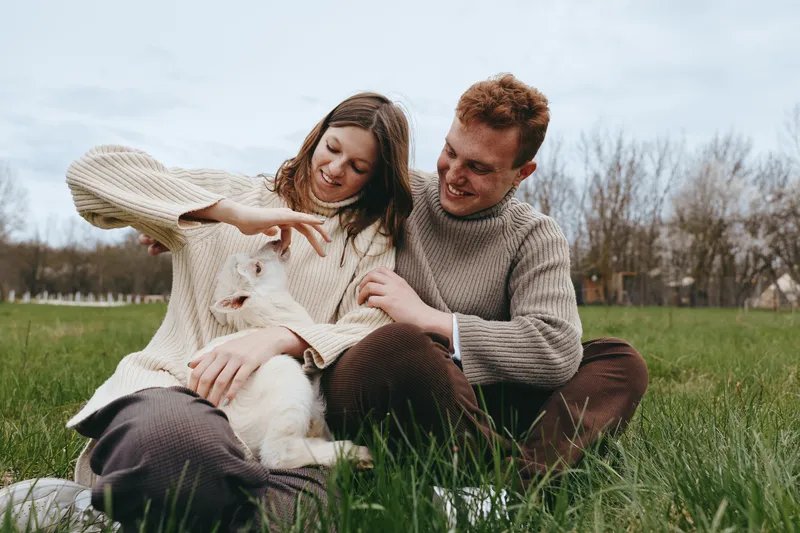 Young couple in warm sweaters plays with a white baby goat sitting on green grass during a calm autumn day in a natural outdoor setting with bare trees in the background.