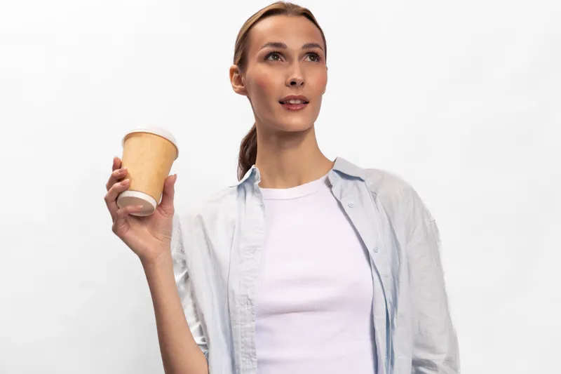 Young woman in casual shirt holding disposable coffee cup looks away thoughtfully isolated on white background. Concept for coffee break, casual lifestyle, and refreshment.