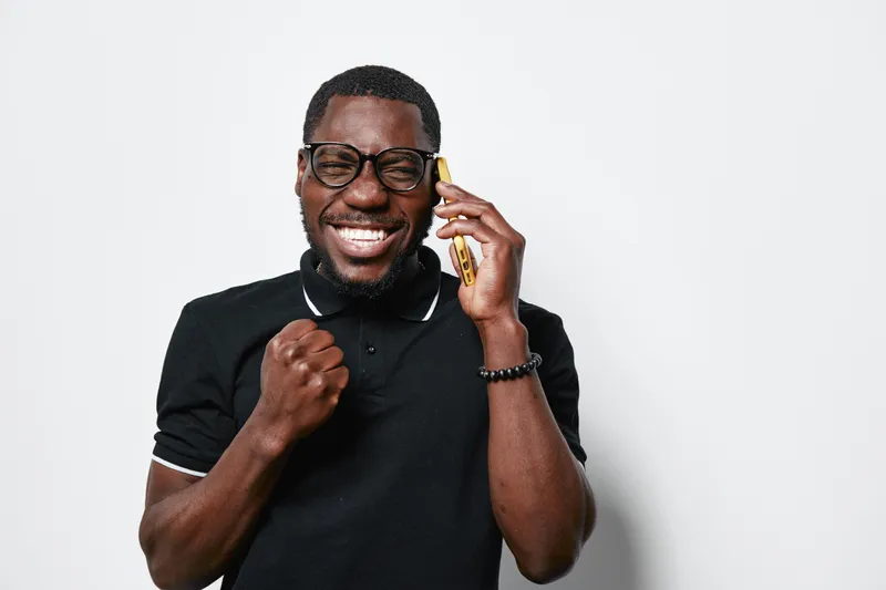 African American man with dark skin wearing glasses and black polo shirt talking on smartphone smiling and celebrating success with fist pump on white background in studio.