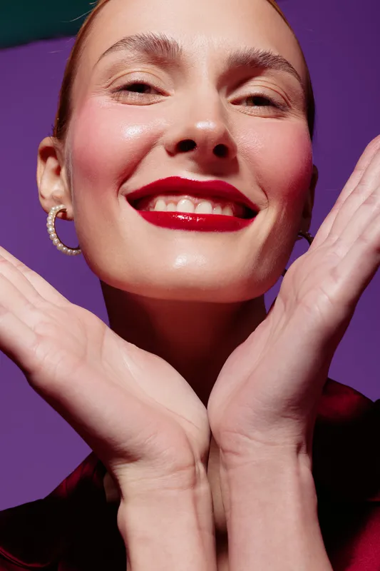 Close-up of smiling woman with red lipstick and pearl earrings posing with hands near face on purple background. Concept of beauty, makeup, skincare, and joyful expression.