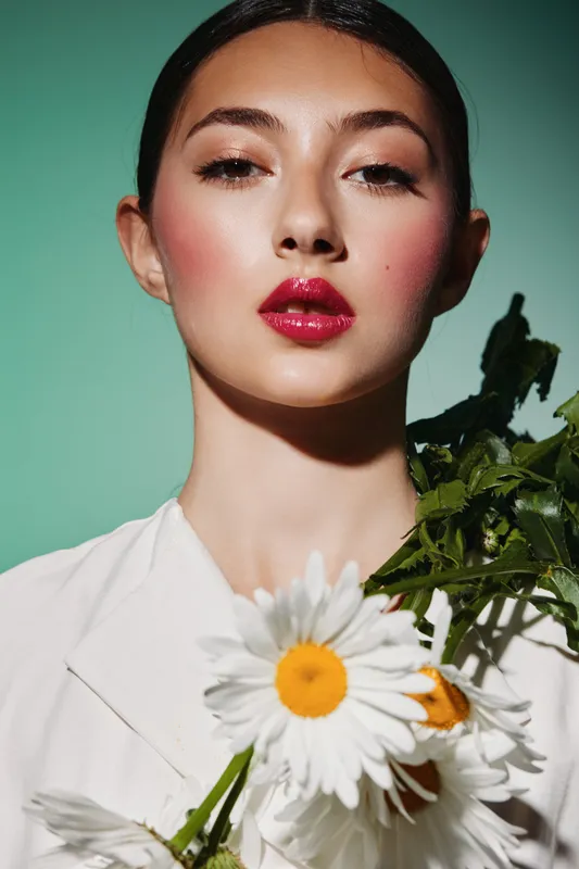 Young woman with glossy red lips and intense makeup holds a bouquet of white daisies against a green background. Fashion beauty portrait for creative and editorial projects.