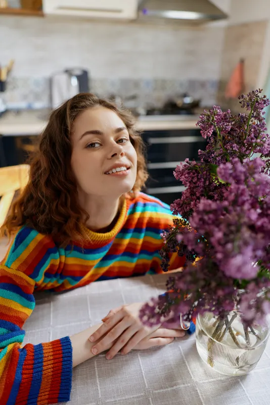 Smiling woman with curly hair leaning on a table in a bright kitchen, wearing a colorful striped sweater, with flowers in the foreground. Lifestyle, home, cozy moment, warm atmosphere.