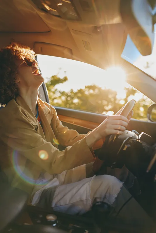 Woman driver enjoying sunlight inside a car, warm golden hour glow on her face, casual attire, peaceful road trip mood, candid lifestyle shot emphasizing travel