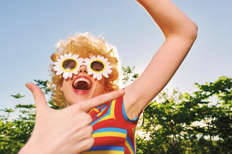 Playful young woman outdoors laughs with flower sunglasses, bright retro vibe and sunlit energy, a carefree moment captured in candid light.