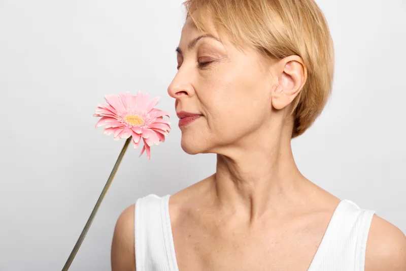 Calm woman holds a pink daisy near her face, eyes closed in profile on a bright background, concept of relaxation and sensory therapy, calm breathing, inner peace
