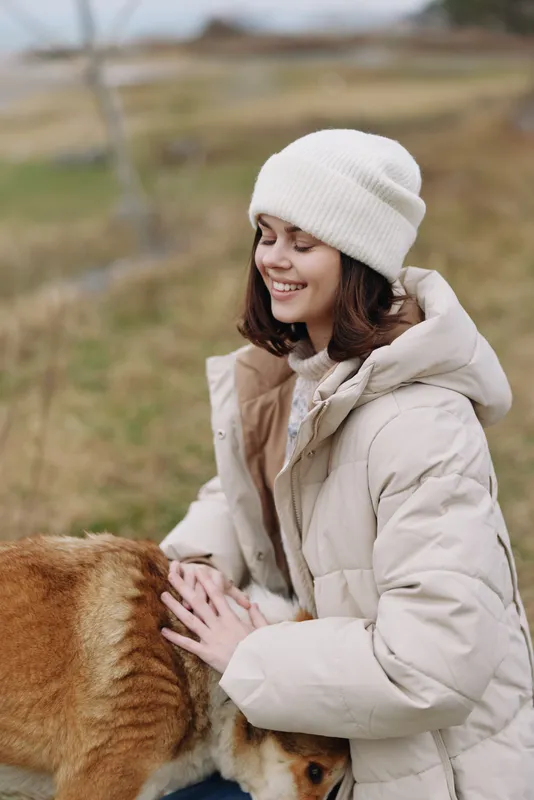 Woman, outdoor scene with a dog, wearing a winter coat and beanie, smiling as she pets a friendly dog in a chilly countryside setting, soft light, cozy mood, calm landscape, natural warmth