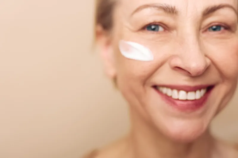Smiling mature woman applies white cream on cheek closeup. Natural skincare concept with healthy skin, wellness, beauty, and facial care in soft warm lighting and beige background.