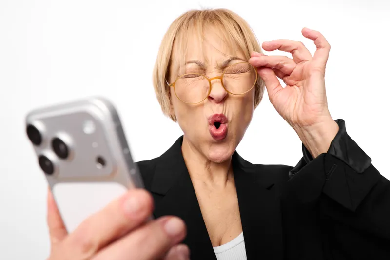 Stressed woman with glasses watches a smartphone, mouth open in anger, holding the device in a business setting, showing frustration on white background today